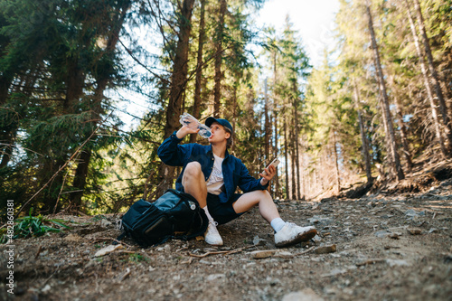 A male tourist in casual clothes sits in the mountains on a trail and sat down to rest, drinking water from a bottle and looking away. Hiker at a halt while climbing a mountain.