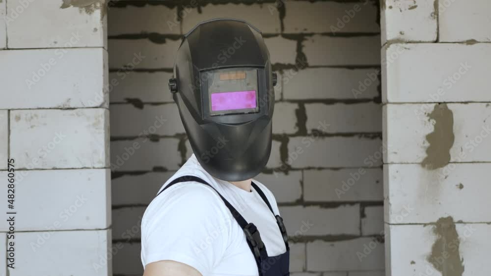 A male construction worker in blue gloves puts on a welding mask against a brick wall A male construction worker in blue gloves puts on a welding mask against a brick wall