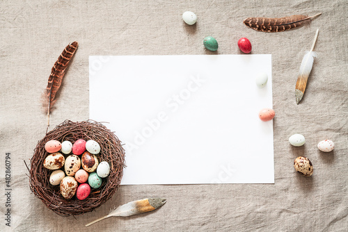 Easter background with white paper blank, colored easter eggs, nest and feathers on a beige background. Easter composition on a textured beige linen background. 