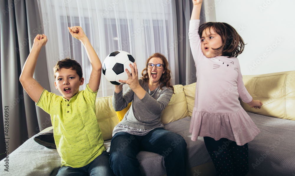 Mother and her kids raising arms and screaming while watching football match and celebrating goal at home Mother and her kids raising arms and screaming while watching football match and celebrating goal at home