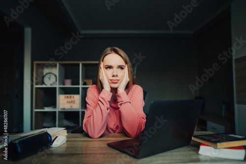 Tired student girl sitting at a table at home with a laptop and books and studying, looking away with a pensive sad face.