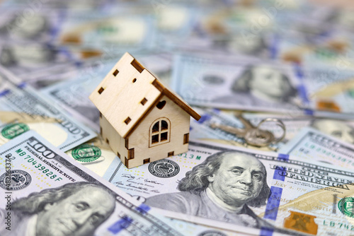 Wooden house model and keys on background of US dollars banknotes. Housing market, purchase or rental of real estate