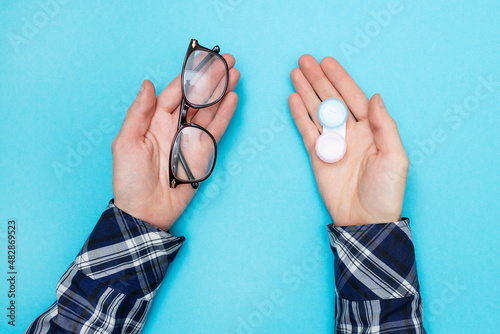 Man holds glasses for vision and container for contact lenses for the purpose of choosing what to use. Blue background. Flat lay.