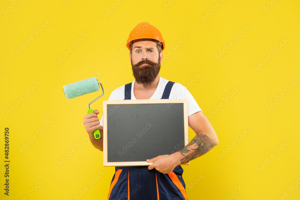 confused bearded man in helmet and work clothes hold paint roller and blackboard confused bearded man in helmet and work clothes hold paint roller and blackboard
