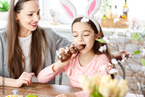 Close up happy mother daughter wearing bunny ears in kitchen during breakfast, eating delicious chocolate rabbit and colorful Easter eggs. Happy Easter atmosphere. Celebrating spring holiday concept.