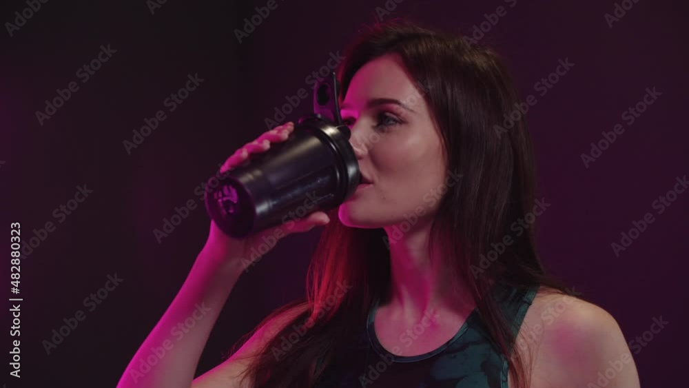 Young pretty woman in sportive top looks in the camera and drinks water from plastic cup in neon lighting Young pretty woman in sportive top looks in the camera and drinks water from plastic cup in neon lighting