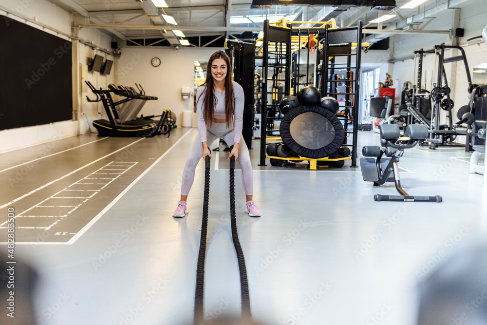 Obraz premium Shot of an attractive young female athlete working out with battle ropes in the gym. Fitness woman working out with battle rope at gym. Beautiful brunette sportswoman doing intense strength training.