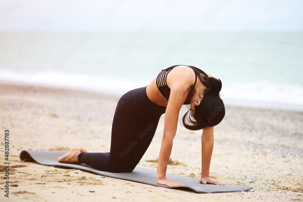 Nice portrait of young beautiful darkhaired woman stretching her back on the seaside. Yoga exercising. Yoga sport. Healthy wellness lifestyle. Spiritual health. Personal fulfillment. - obrazy, fototapety, plakaty 