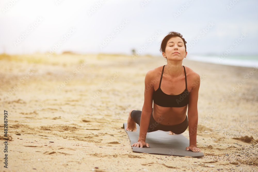 A portrait of beautiful woman standing in plank position and stretching her back on the beach. Yoga sport. Healthy wellness lifestyle. Spiritual health. Personal fulfillment. - obrazy, fototapety, plakaty 