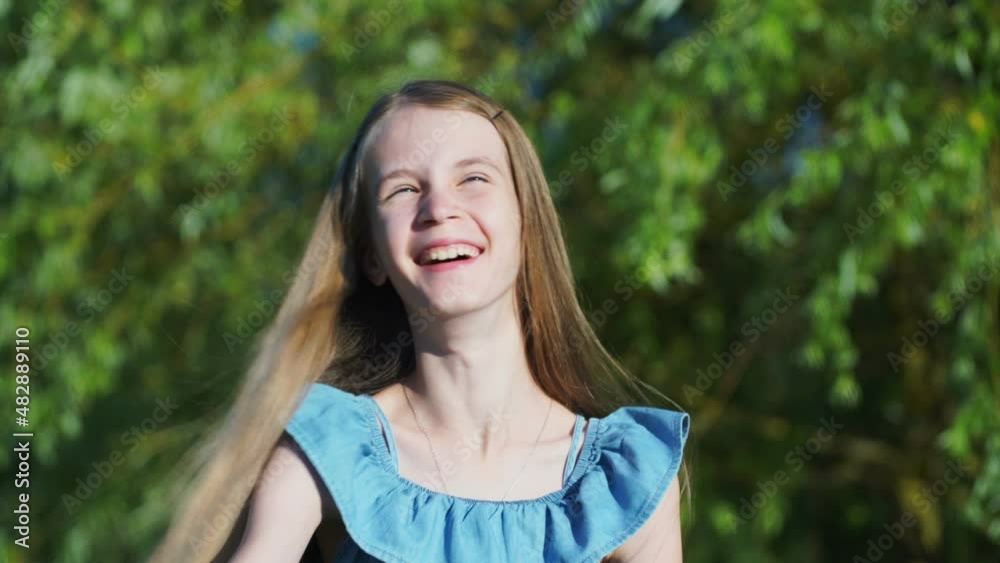 Cute teenage girl in stylish blue dress laughs and adjusts blonde hair standing against green trees in sunny summer garden Cute teenage girl in stylish blue dress laughs and adjusts blonde hair standing against green trees in sunny summer garden