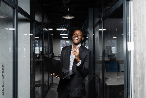 Cheerful black business manager with laptop is pointing fingers up and smiling in office.