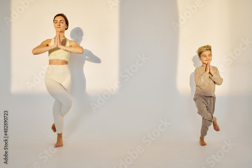 brunette woman with her blonde child son making yoga poses on white background, sunset light in studio