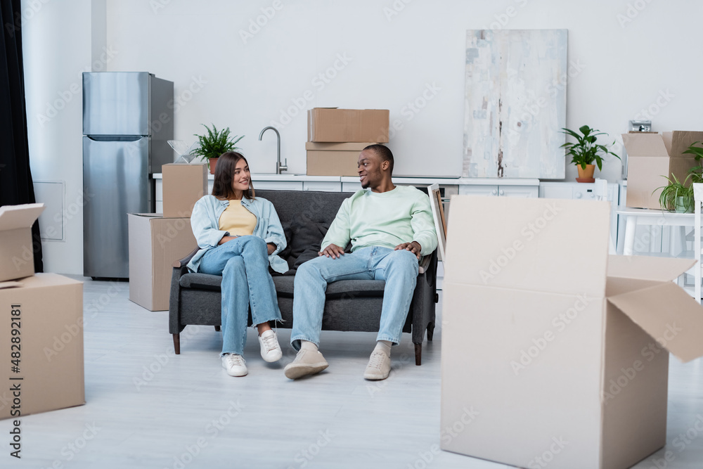 happy interracial couple sitting on sofa around carton boxes during relocation. happy interracial couple sitting on sofa around carton boxes during relocation.