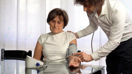Fototapeta Naklejka Na Ścianę i Meble -  70 year old old lady woman tests blood pressure at home - private doctor visit in apartment  during Covid-19 Coronavirus lockdown epidemic