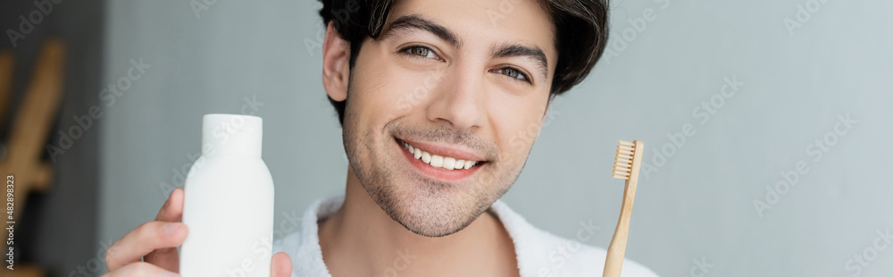 Fototapeta premium cheerful man holding toothpaste and toothbrush while looking at camera, banner.