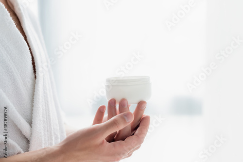 cropped view of man in bathrobe holding container with cosmetic cream.
