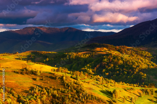 mountainous rural landscape at cloudy sunset. forested slopes with rural fields in evening light. village in the distant valley behind the hill. ridge with snow stripes on the summit in the distance