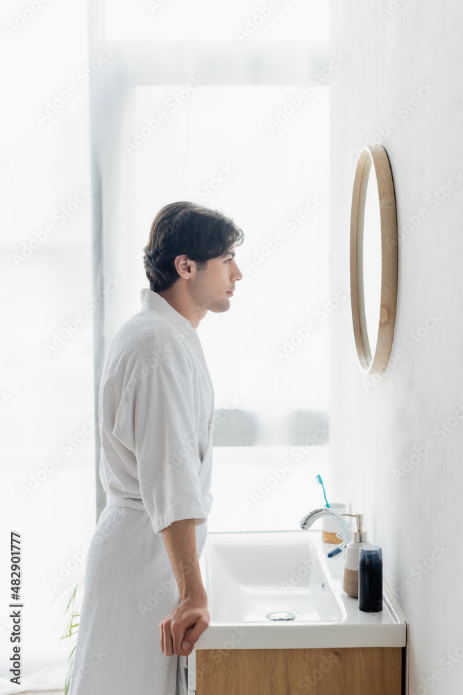 side view of man in bathrobe looking in mirror near toiletries on sink.