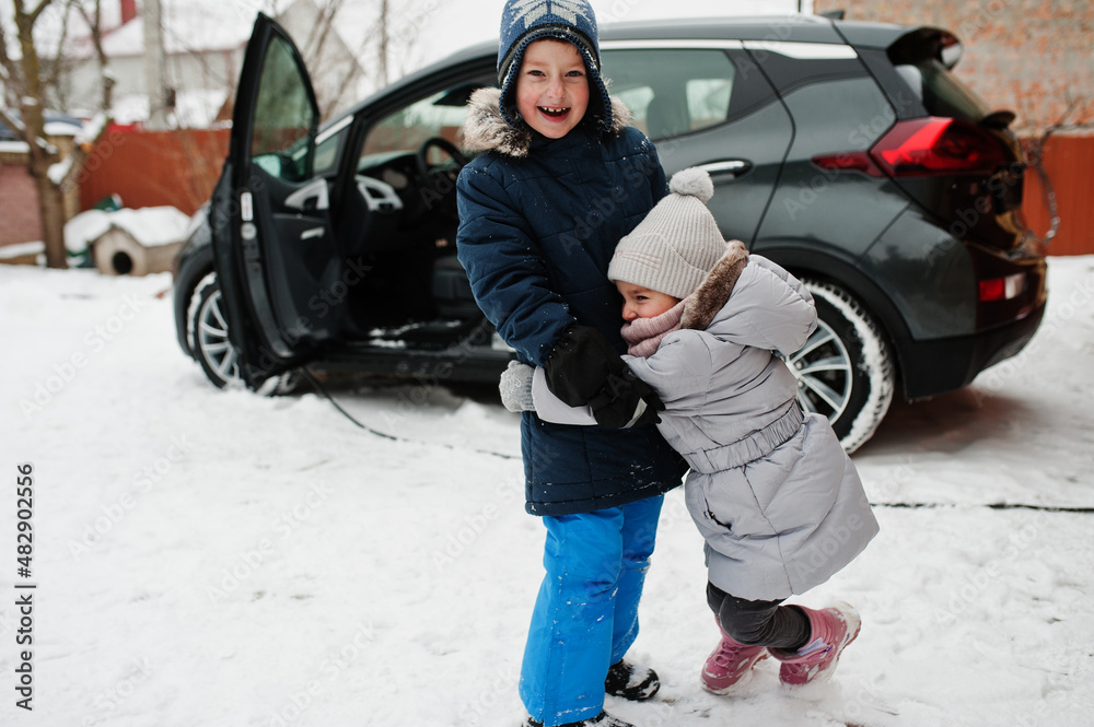 Brother with sister play against charging electric car in the yard of house at winter.