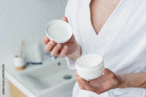 cropped view of man in white bathrobe holding cosmetic cream in blurred bathroom.