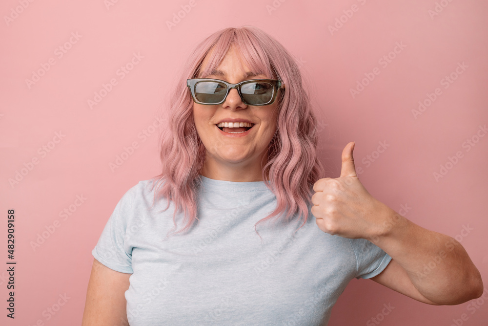 Happy young caucasian woman in bright stylish sunglasses and a blue t-shirt showing thumbs up and smiling isolated over pink background. Happy young caucasian woman in bright stylish sunglasses and a blue t-shirt showing thumbs up and smiling isolated over pink background.