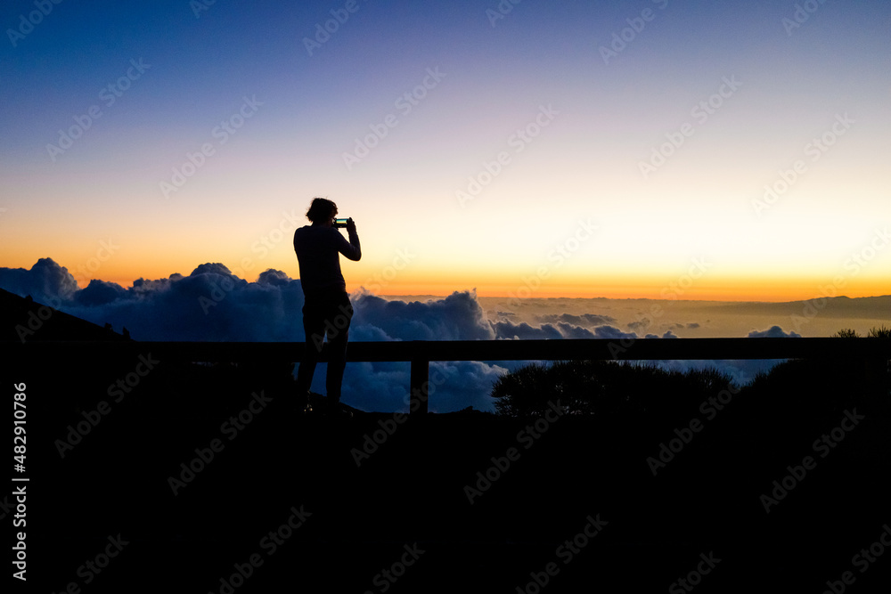 Silhouette of a woman photographs a sunset on top of a mountain above the clouds