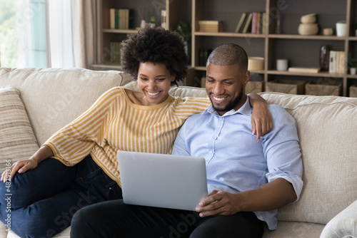 Beautiful cheerful African boyfriend and girlfriends relax on sofa with laptop, put wireless computer on laps watching streaming digital online TV, smiling enjoy video vlog, using e-services concept