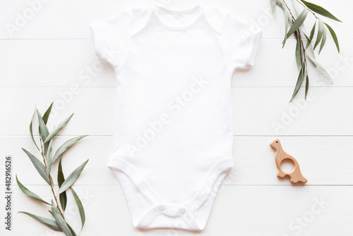 Mockup of white baby bodysuit on wood background with greenery and toy. Blank baby clothes template, flat lay.