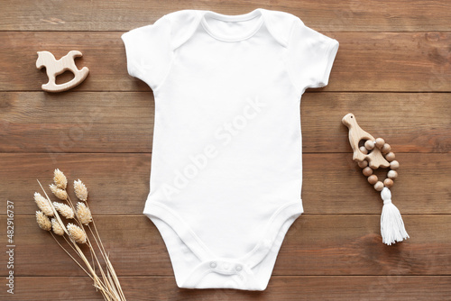 Mockup of white baby bodysuit on dark wood background with dried flowers and toys. Blank baby clothes template, flat lay. photo