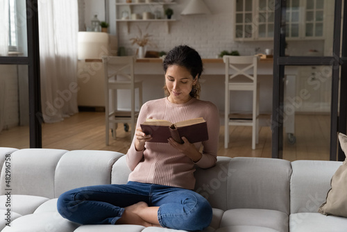 Young 30s Hispanic woman holding paper book seated cross-legged on sofa at home, reading exciting bestseller literature, enjoy lazy weekend, day off free time with favourite hobby. Leisure concept #482916758