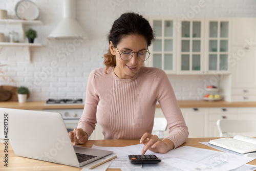 Hispanic woman use calculator calculate costs working sit at table in kitchen. Pay monthly bills through e-bank app on laptop. Business, summarize household expenses, manage budget, accounting concept © fizkes