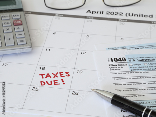 A 2022 calendar noting the April 18 USA Internal Revenue Service IRS income filing deadline for year 2021 taxes is shown up close, with a calculator, ink pen, eye glasses, and Form 1040 paperwork.