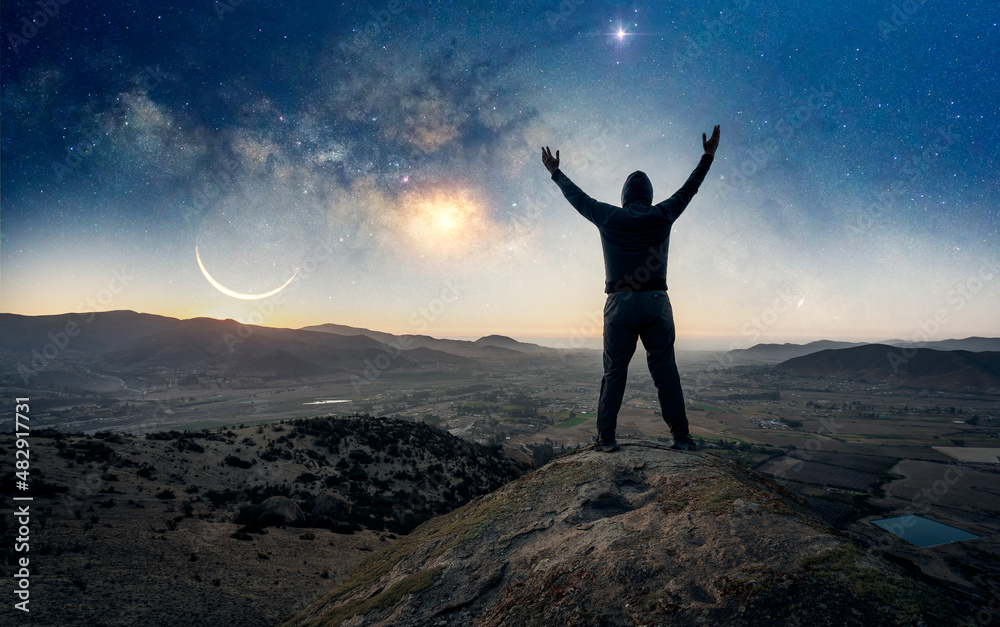 person silhouette standing on the top of the mountain, back view, with hands up celebrating under the Milky Way
