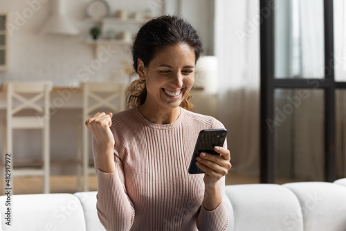 Excited 30s Latina woman sit on sofa with smartphone read great news feels happy look overjoyed got unbelievable offer. Lottery win, got hired job of dream, celebrate moment of auction victory concept photo