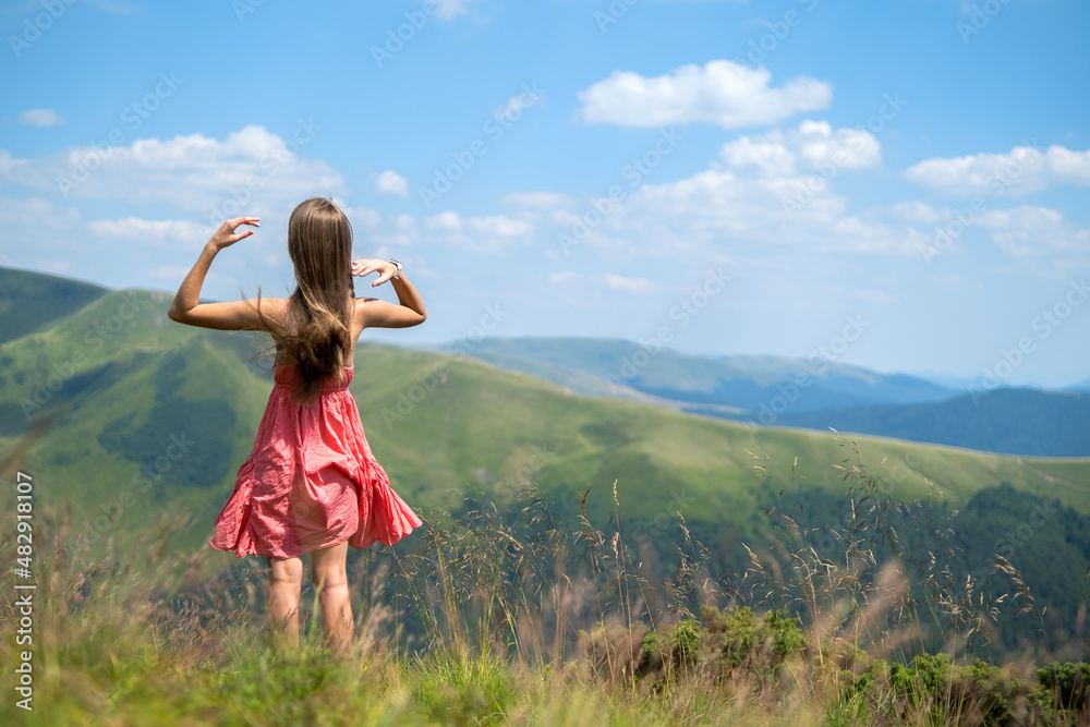 Young happy woman traveler in red dress standing on green grassy hillside on a windy day in summer mountains enjoying view of nature Young happy woman traveler in red dress standing on green grassy hillside on a windy day in summer mountains enjoying view of nature
