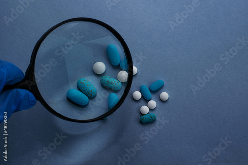 Doctor's workplace. Top view on a light background with scattered pills. A doctor in blue medical gloves holds a magnifying glass in his hand. Medical concept.