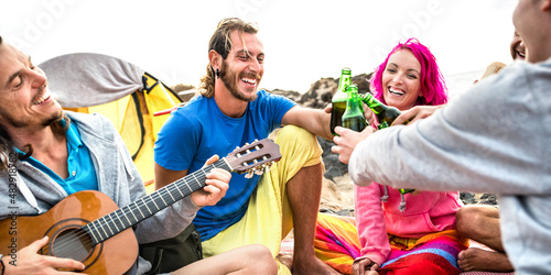 Hippie friends having fun together at beach camping party - Life style travel concept with young hipster travelers playing guitar and drinking bottled beer at summer surf camp - Warm vivid filter