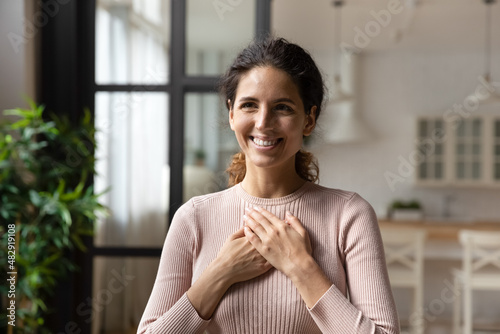 Head shot portrait young attractive Hispanic woman standing alone at home put folded palms on chest feels grateful, express appreciation, sincere feelings smiles looks aside. Believe, charity concept photo