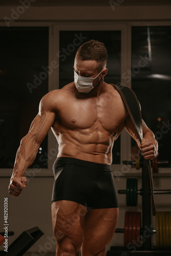 A shirtless bodybuilder in a medical face mask is posing after a workout in gym