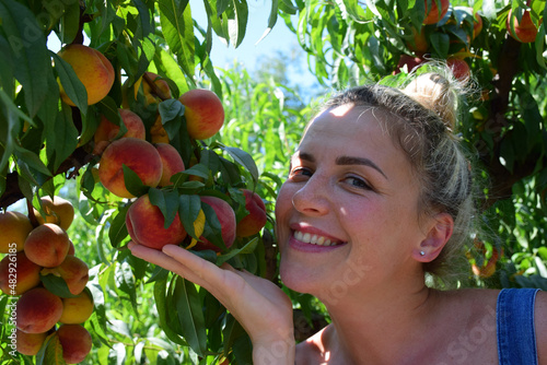 Attractive blonde girl holds a peach tree branch with fruits with her hand and smiles