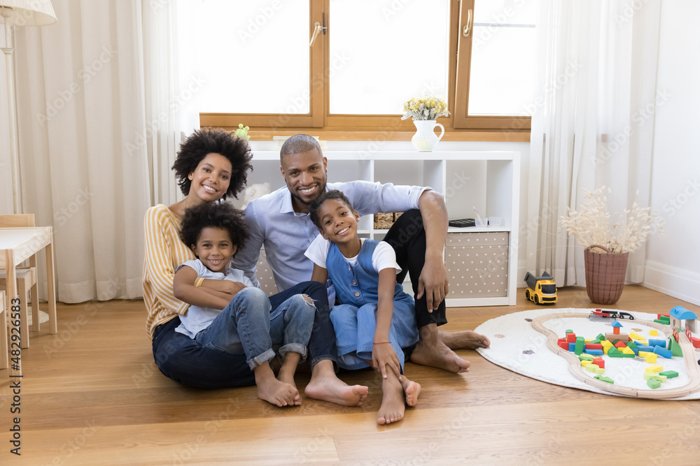 Young African married couple sit on warm floor in cozy playroom hugging their little cute children smiling looking at camera. Happy homeowners family portrait, bank loan, tenancy, new house concept Young African married couple sit on warm floor in cozy playroom hugging their little cute children smiling looking at camera. Happy homeowners family portrait, bank loan, tenancy, new house concept