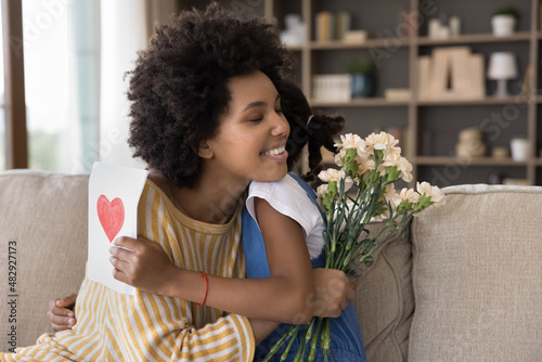 Loving African daughter congratulates young mom on birthday, mum receive flowers, handmade postcard on International Women Day, heart symbol of love and family ties. Happy Mother Day congrats concept