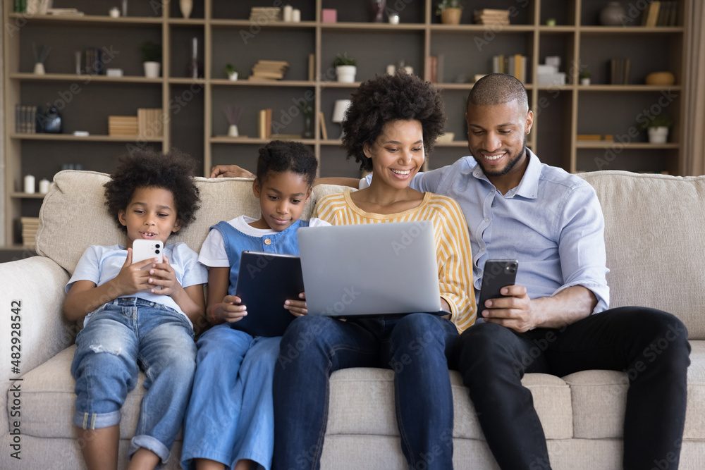 African spouses and children sit on sofa enjoy leisure use diverse electronic gadgets. Kids absorbed in videogame, instead live communication prefer online fun, bad life habit, devices overuse concept African spouses and children sit on sofa enjoy leisure use diverse electronic gadgets. Kids absorbed in videogame, instead live communication prefer online fun, bad life habit, devices overuse concept