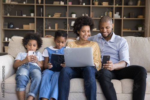 African spouses and children sit on sofa enjoy leisure use diverse electronic gadgets. Kids absorbed in videogame, instead live communication prefer online fun, bad life habit, devices overuse concept