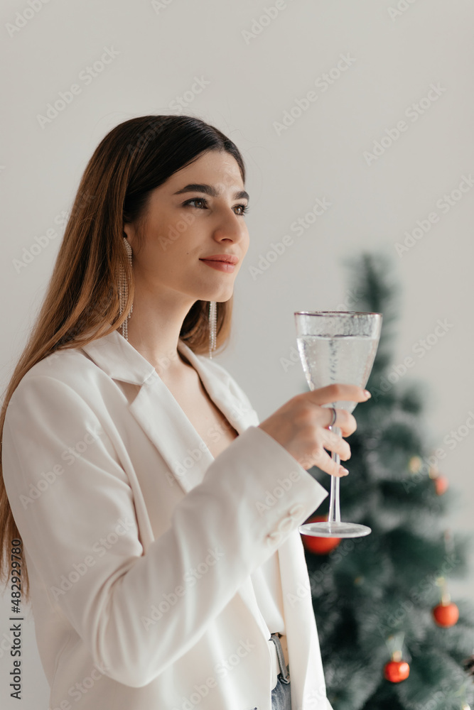 A young pretty girl in a white jacket and earrings with brown long hair smiles with a glass of champagne against the background of a festive Christmas tree and a white wall A young pretty girl in a white jacket and earrings with brown long hair smiles with a glass of champagne against the background of a festive Christmas tree and a white wall