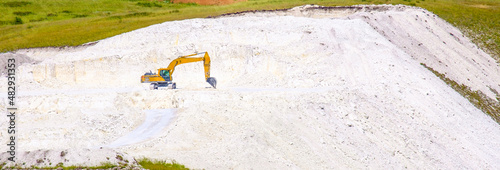 A yellow excavator digs stone chalk in an open pit on a summer day. Widescreen image. Banner.