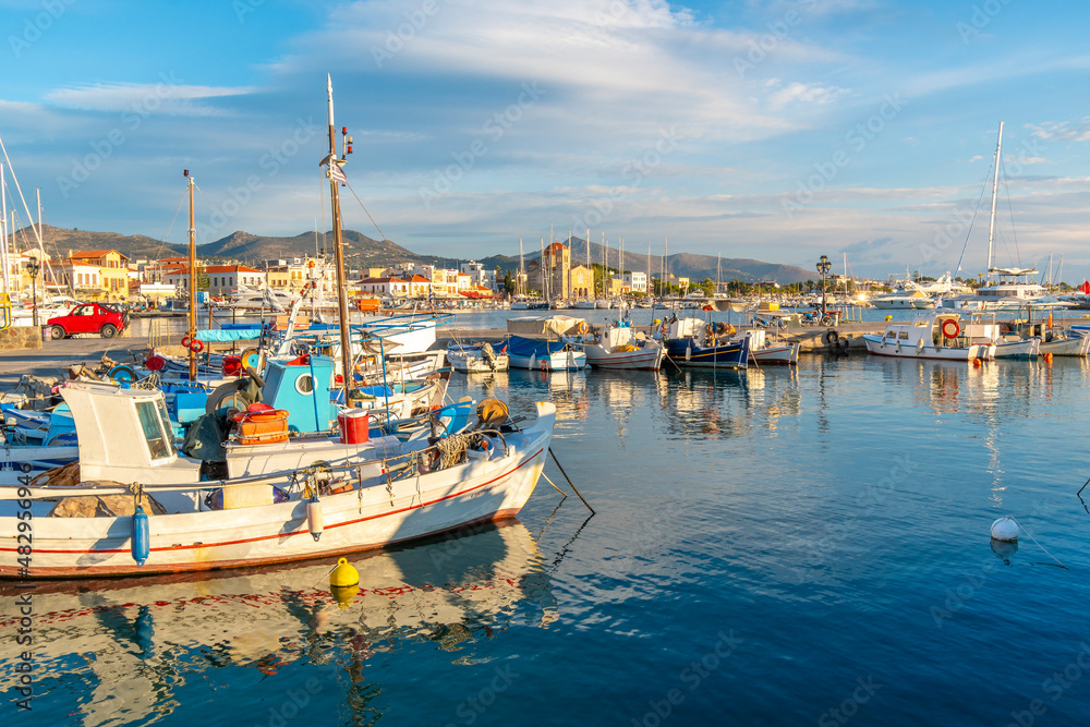 Colorful fishing boats line the harbor of the Greek island of Aegina, Greece at dusk, with the waterfront promenade and village in view.	