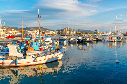Colorful fishing boats line the harbor of the Greek island of Aegina, Greece at dusk, with the waterfront promenade and village in view. 