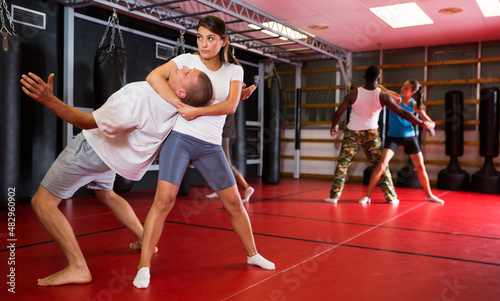 Woman practising neck grabbing self-protective move with man during group training. African-american man sparring with woman in background.