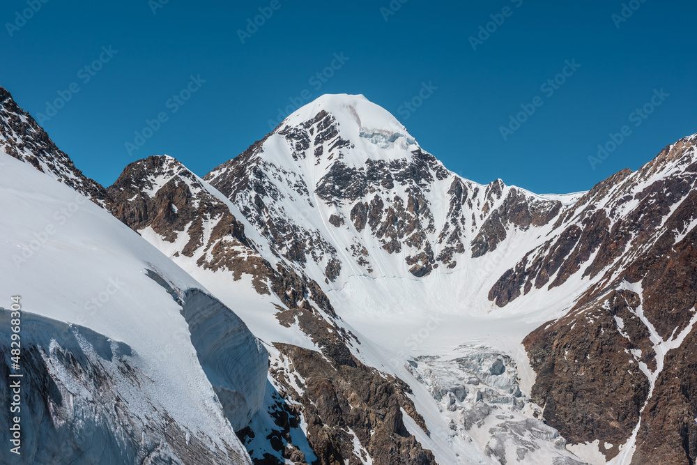 Scenic alpine landscape with snow-covered mountain top and steep glacier tongue with icefall in sunlight. Awesome mountain scenery with pointed peak and glacier on rocks under blue sky in sunny day.